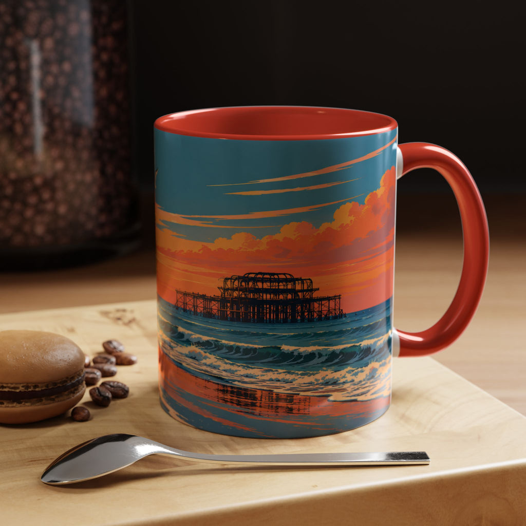 A ceramic coffee mug with a red handle and interior, featuring a colorful printed illustration of Brighton Pier at sunset. The mug is set on a wooden table next to a spoon and a macaron, with a dark background.
