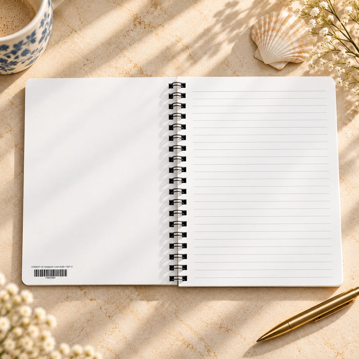 An open white spiral-bound lined notebook lies on a light beige stone surface with soft shadows. A blue-and-white ceramic cup is partially visible at the top left, a seashell at the top right, and a gold pen at the bottom right.
