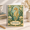 A spiral-bound notebook with an ornate vintage-style cover featuring a seahorse and decorative floral motifs in teal, green, cream, and gold tones. The notebook stands on a tabletop with a cup, books, a pen, and dried flowers in the background.